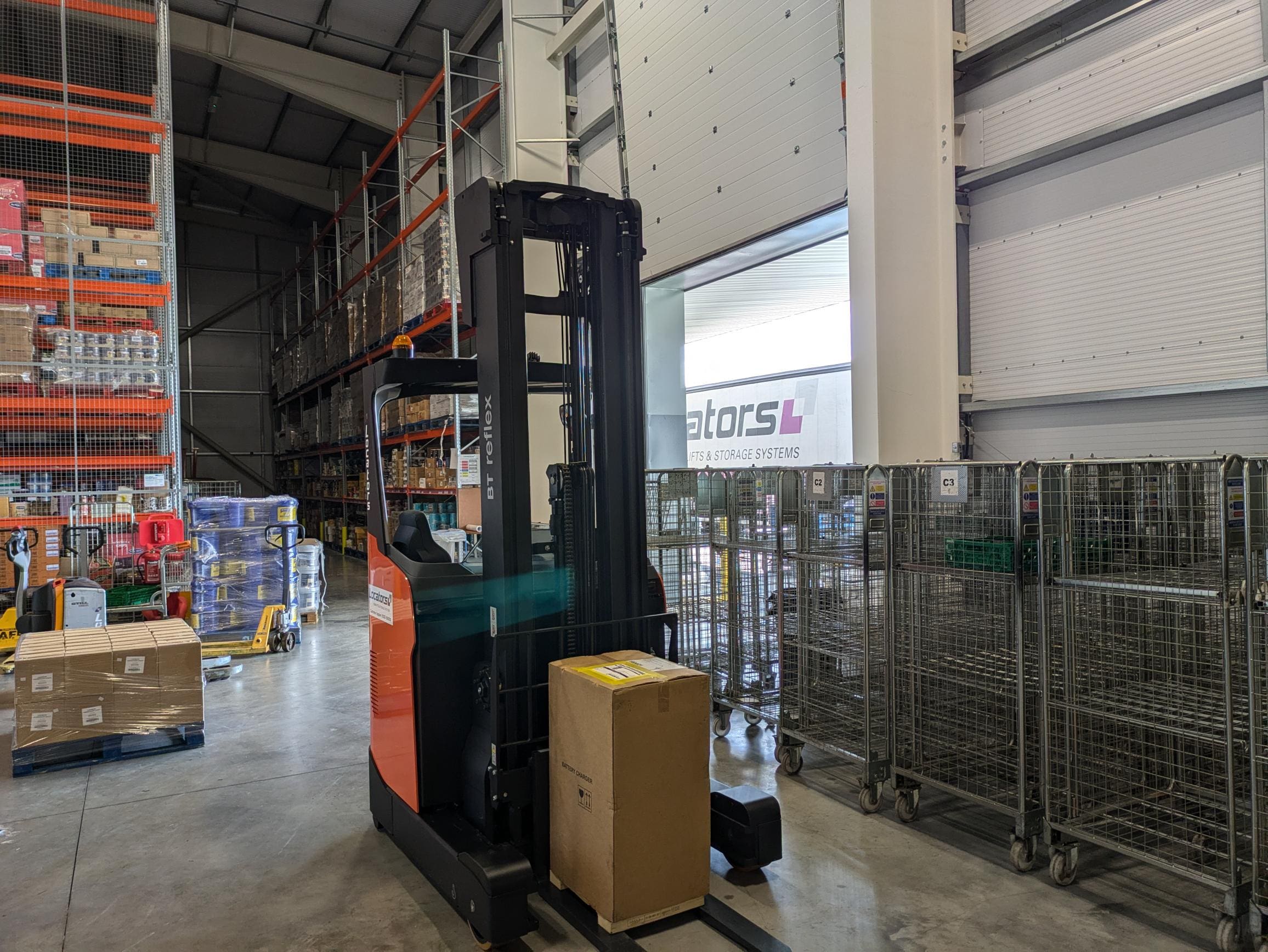 Warehouse with an orange and black Toyota BT Reflex forklift carrying a box, surrounded by metal racks filled with various products and equipment. The Locators logo is visible in the background.