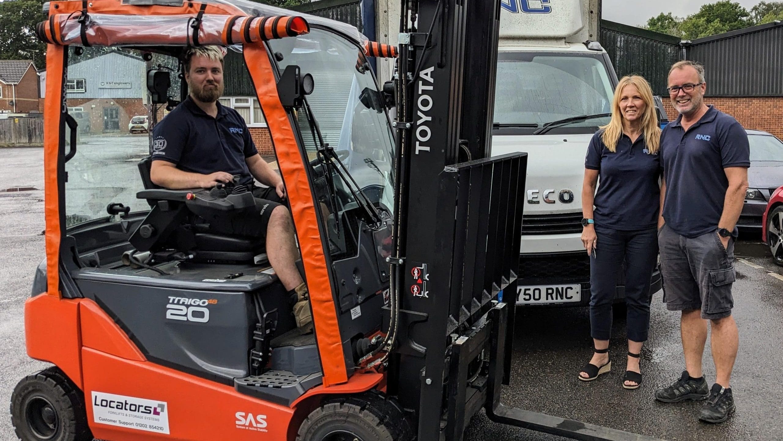 three individuals with a forklift, prominently labeled with the Locators logo, highlighting their forklift hire services in Hampshire. A male employee is seated in the driver's seat of a bright orange Toyota Traigo 20 forklift, demonstrating readiness for operation. Beside the forklift, a woman and another man, both wearing dark blue Locators uniforms, stand smiling, suggesting a team-oriented, customer-friendly service.